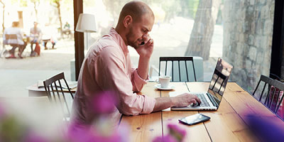  Man sitting at a desk while browsing his laptop and talking on a cellphone. 