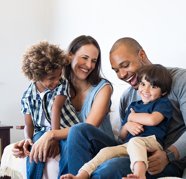 A happy father, mother, and two young boys sit on a couch enjoying time together