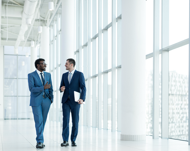 Two men in business attire are walking and talking while inside an office building.