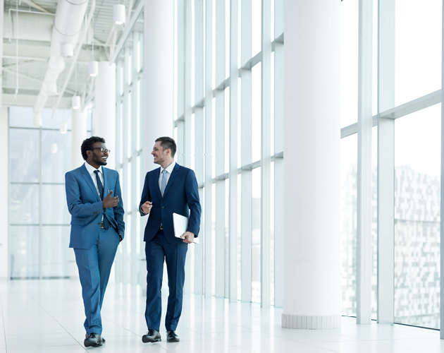 Two men in business attire are walking and talking while inside an office building. 