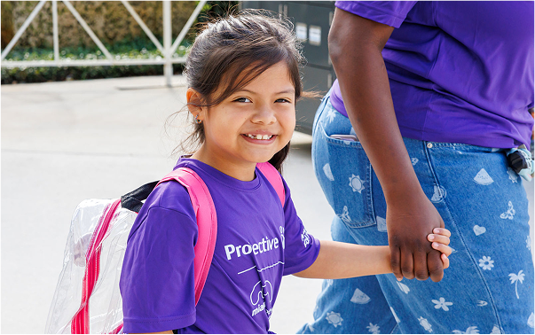 A girl wearing a shirt with the Protective logo and a backpack holds hands with a caregiver.