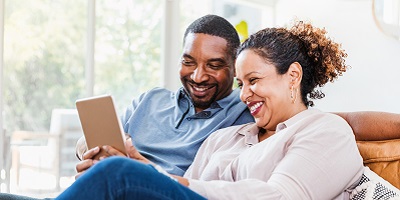 African American couple sitting on the couch, laughing while looking on tablet together