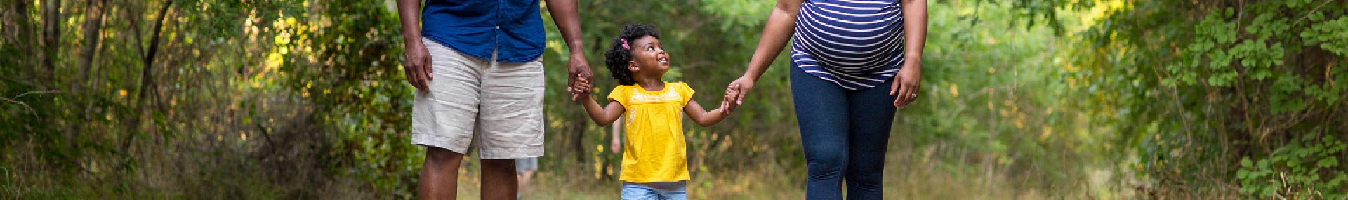 Father and pregnant mother walk with their daughter through a wooded area holding hands.