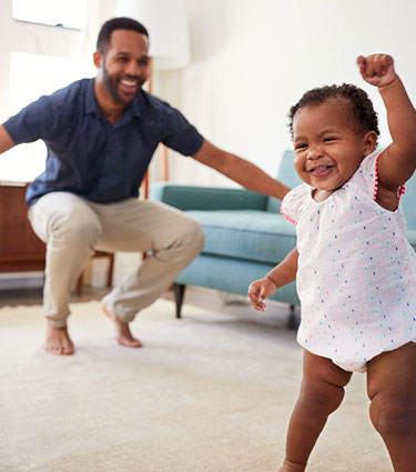 Infant girl taking first steps with dad celebrating in background