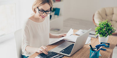 Woman in glasses, smiling, doing work at a desk. 