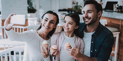 Smiling mom holds out her phone to take a photo of herself with husband and young daughter posing with pretty cupcakes at a bakery