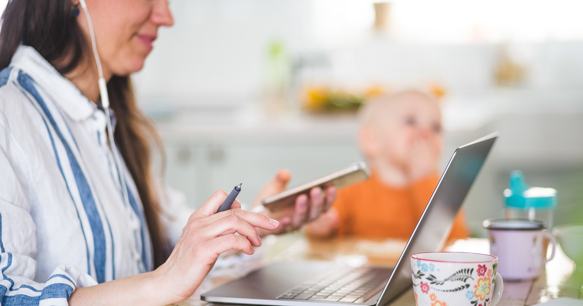 Woman wearing earbuds using a smartphone and laptop at kitchen table with baby in a highchair next to her.