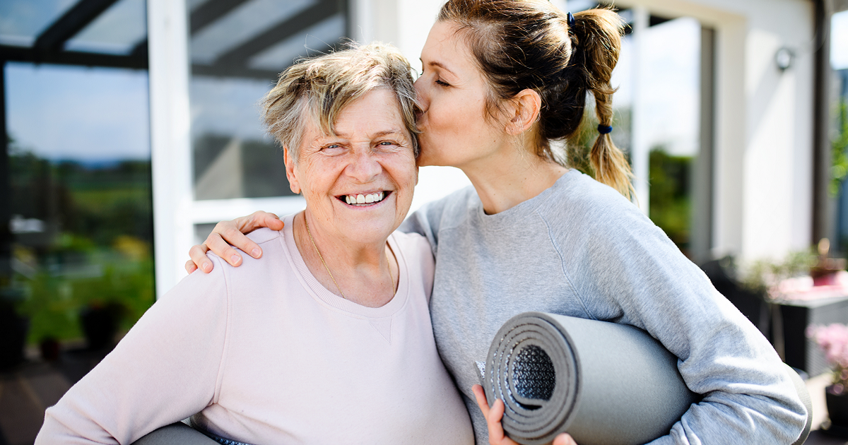 Woman holding yoga mat under arm with other arm around elderly mother and kissing her on the cheek