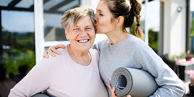 Woman holding yoga mat under arm with other arm around elderly mother and kissing her on the cheek