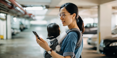 A mother wearing a baby holder while browsing her phone for life insurance policies.