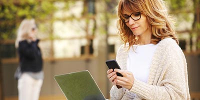 Woman in glasses sitting outside looking at her phone with her laptop in her lap.