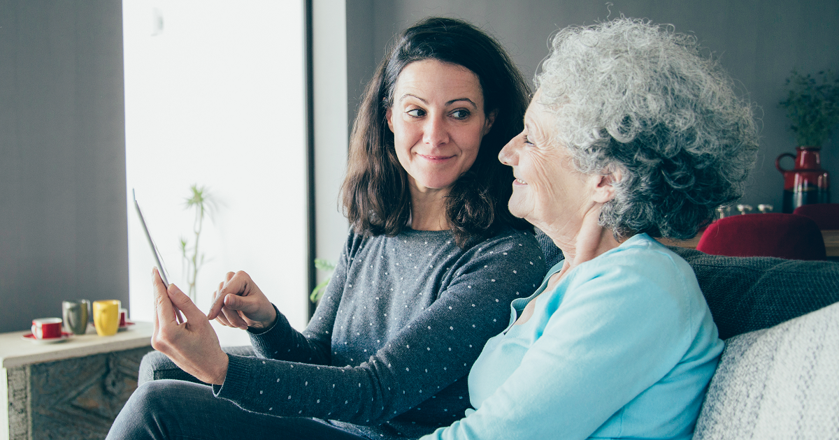 Woman sitting on couch with elderly mother and using a tablet.