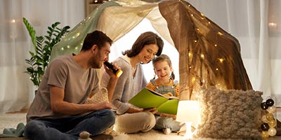 Mom, dad and their young daughter camping in a tent in the living room and reading a book.