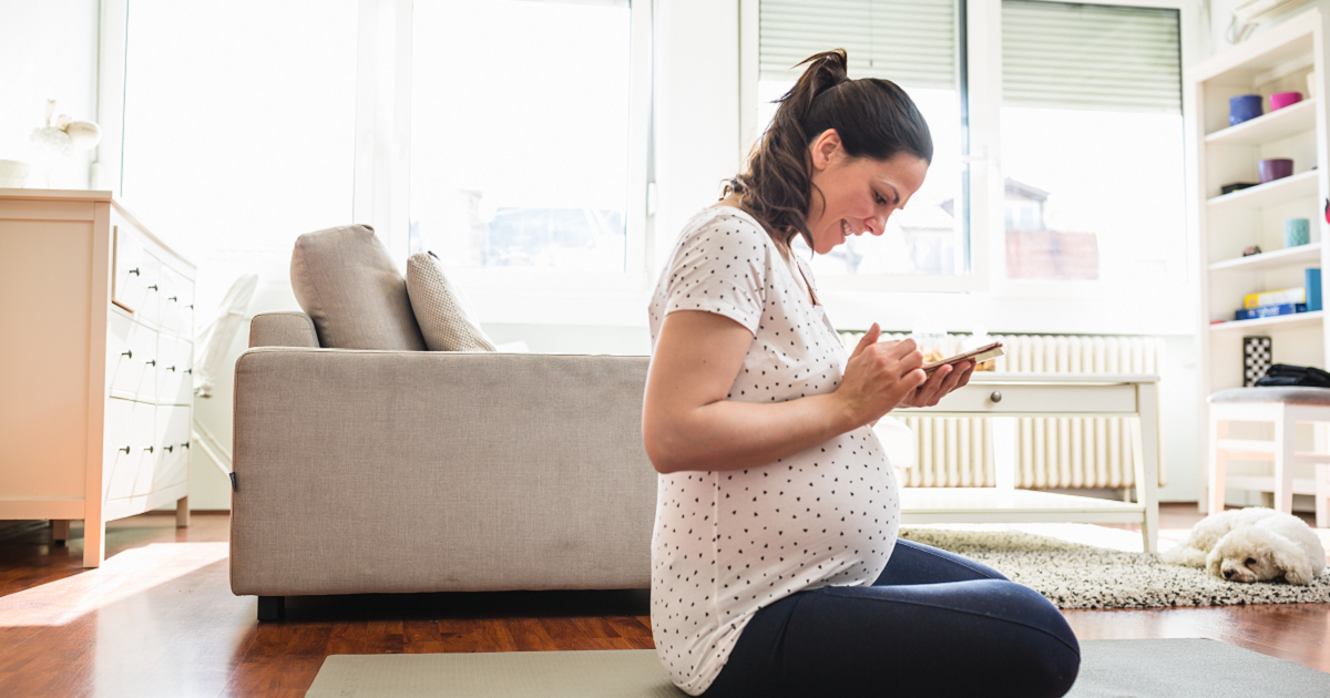 Pregnant woman sitting on yoga mat in living room looking at smartphone with dog on floor beside her