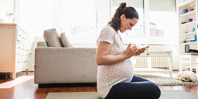 Pregnant woman sitting on yoga mat in living room looking at smartphone with dog on floor beside her