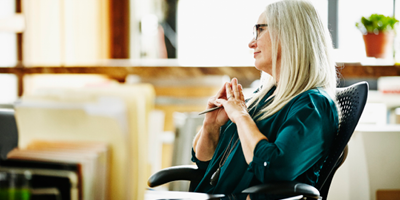 A woman in a dark green shirt leans back in her chair and smiles at work.