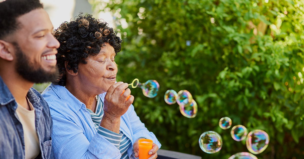 Adult son sitting outside next to elderly mom as she blows bubbles