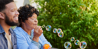 Adult son sitting outside next to elderly mom as she blows bubbles