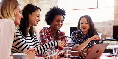 Four women in their thirties, sitting at a coffee bar looking at a computer and smiling.