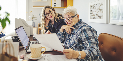 Senior woman and younger woman sitting at desk in front of laptop looking at estate papers