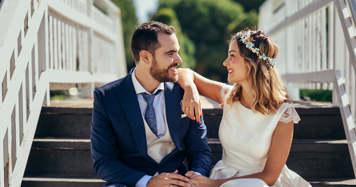 Bride and groom sit outside on stairs holding hands and looking at each other.
