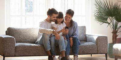 Picture of mom and dad sitting on the living room couch with their young daughter in-between, looking at a mobile phone.