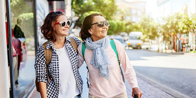 Grandmother and college-age granddaughter walking down city street wearing sunglasses and backpacks 