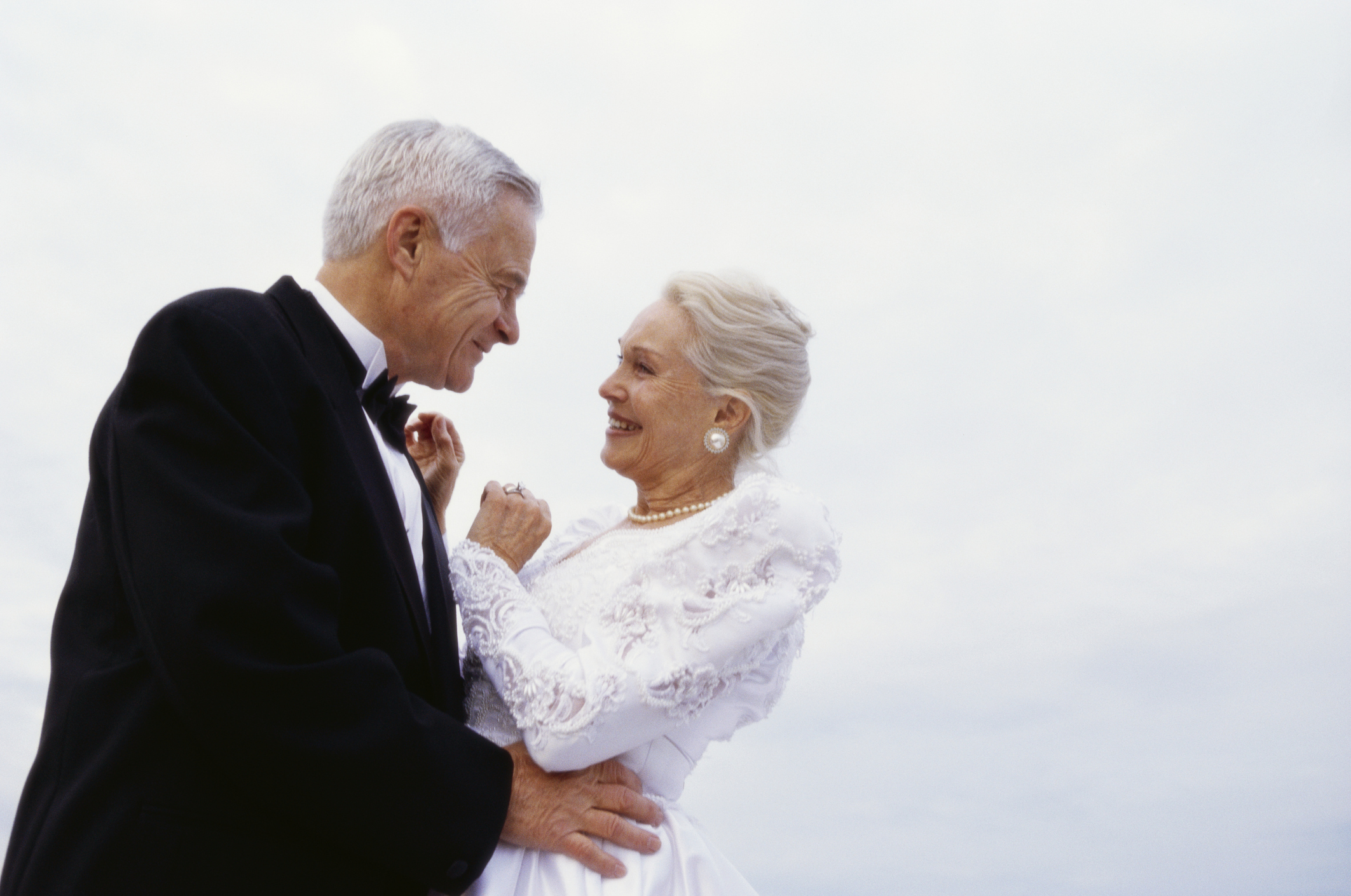 A newlywed senior couple on the beach in their wedding outfits smiling lovingly at each as they dance.