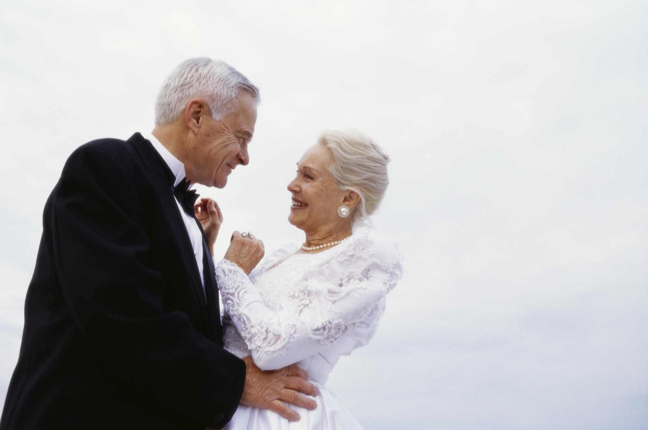 A newlywed senior couple on the beach in their wedding outfits smiling lovingly at each as they dance. 