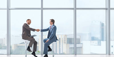 Two businessmen on tall stools, shaking hands in front of the window of an office building.
