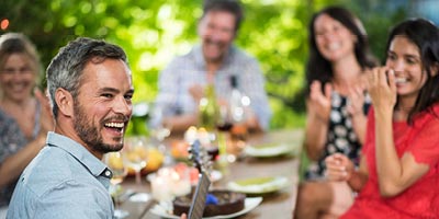  Smiling middle-aged man plays guitar and laughs with friends around table at restaurant