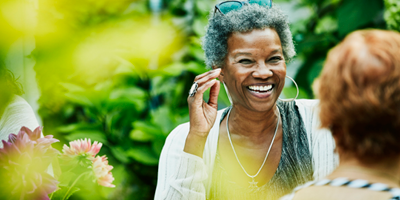 Two women happily engage in conversation in an outdoor setting with greenery.