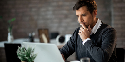 Man in a gray sweater thoughtfully looking at his laptop, as if studying information.