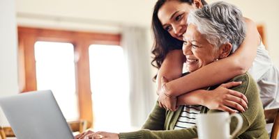 Adult woman smiling, standing behind her mom with her arms around her as they look at a laptop together.