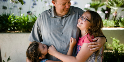 Man embracing young daughters in an outdoor setting.
