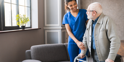 A young nurse in blue scrubs helps an elderly man using a walker.