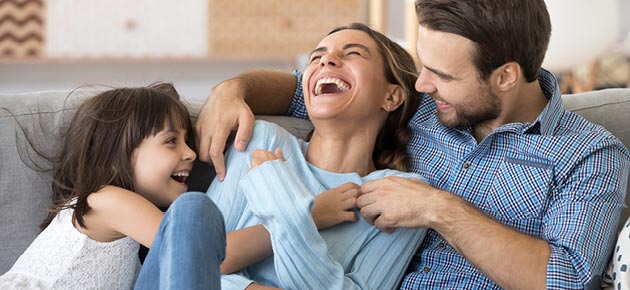 Young family of three on a couch. Child and Dad are tickling mom and she is laughing.  